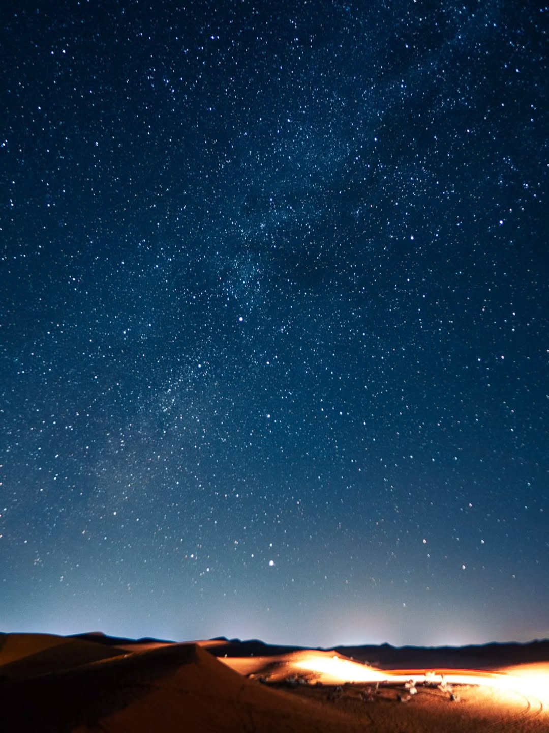 Milky Way over the Sahara dunes with the warm glow of a desert camp on the horizon