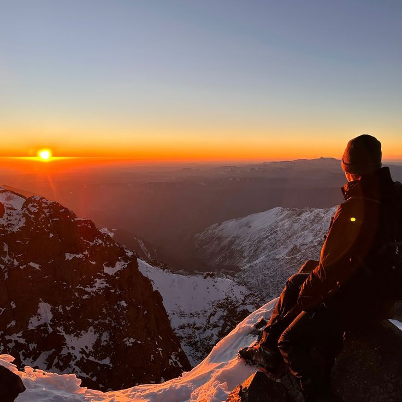 A trekker watching sunrise over snow-covered High Atlas ridges from near the Toubkal summit